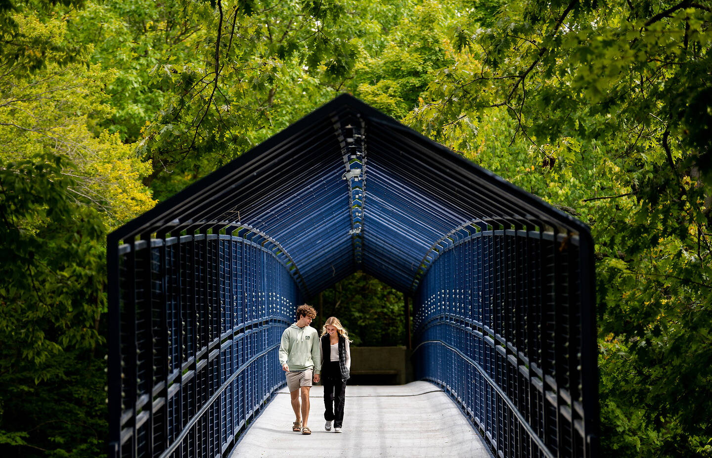 Two people walk hand-in-hand across a blue metal pedestrian bridge surrounded by lush green trees.